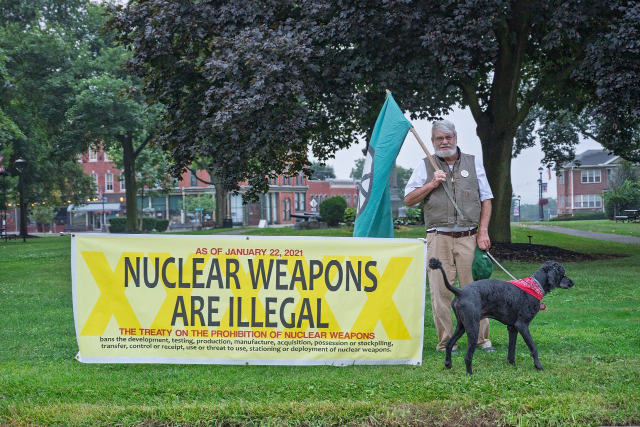 A man holding a green flag stands next to a yellow banner that reads 'Nuclear Weapons Are Illegal' in a park, alongside a dog wearing a red bandana.